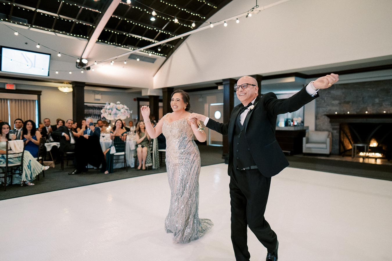 A joyful couple dancing at their wedding reception, surrounded by guests seated at elegantly decorated tables under twinkling string lights in a spacious banquet hall.
