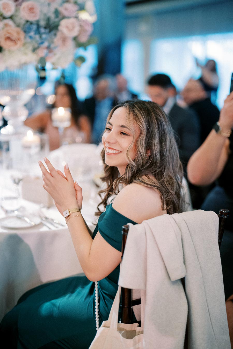 A woman in a green dress smiles and claps at an elegant event, seated at a decorated table with candles and floral arrangements.