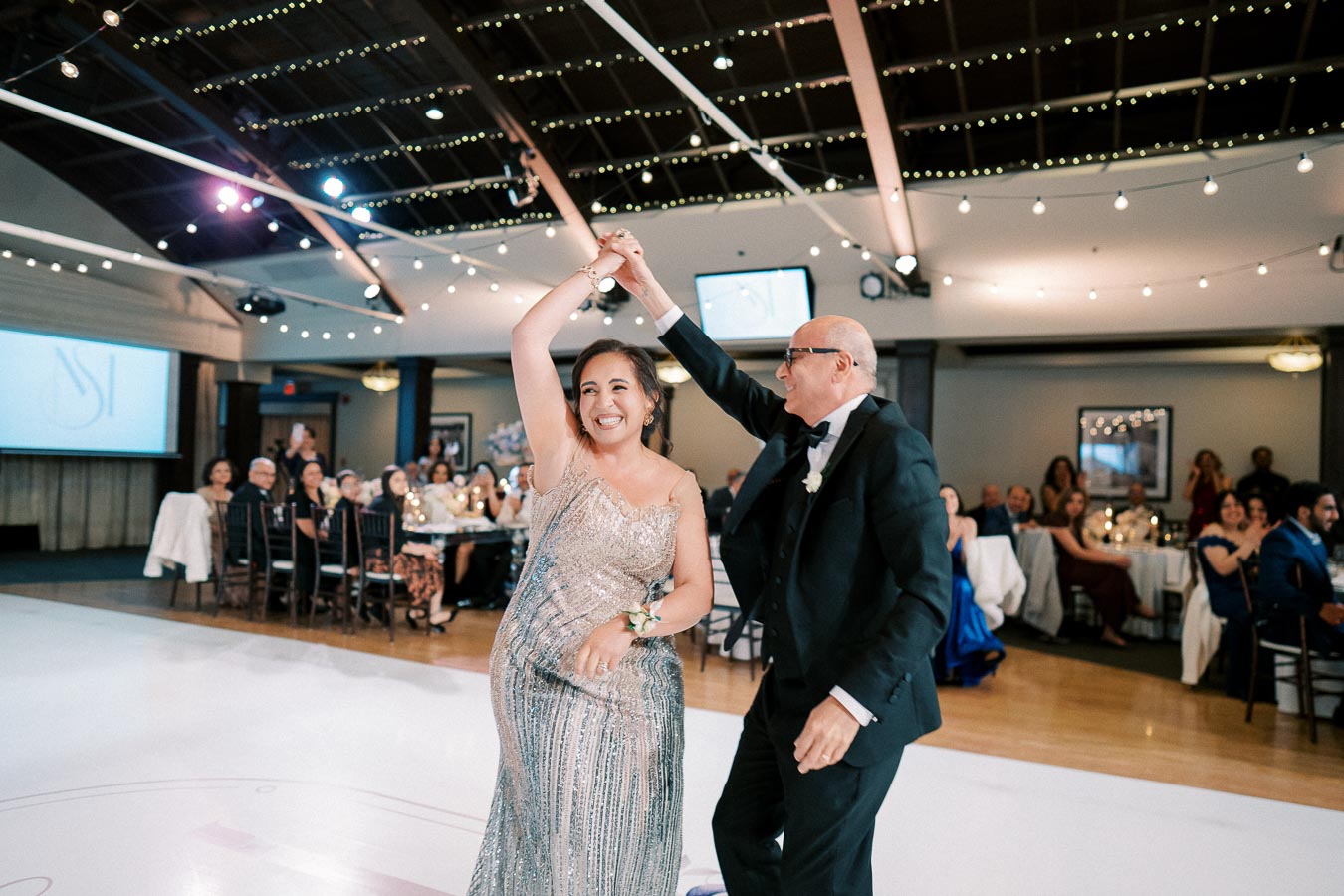 A joyful couple dancing at a wedding reception, with twinkling string lights and elegantly dressed guests in the background.