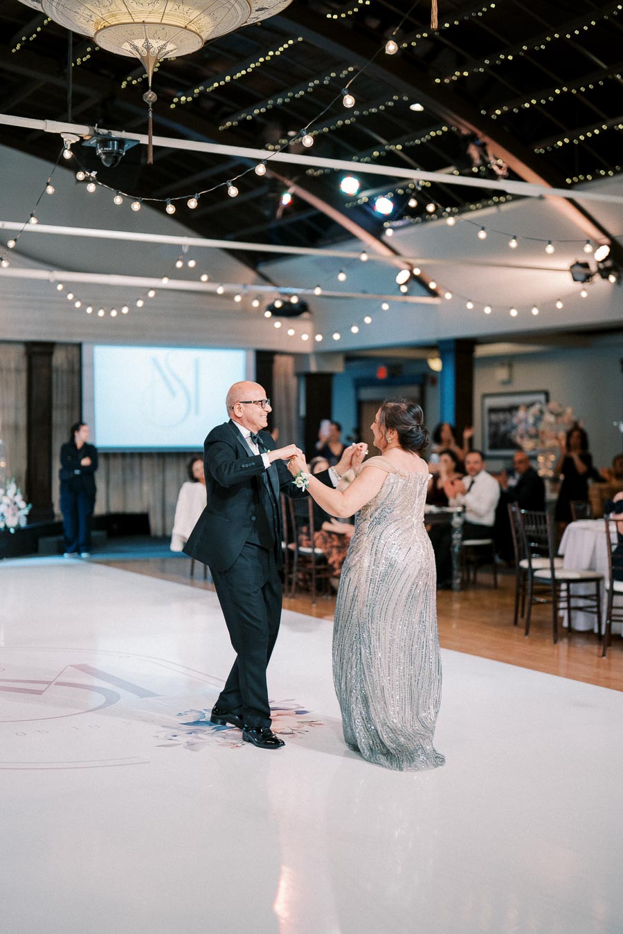 Elderly couple dancing joyfully at a wedding reception in an elegantly decorated venue with string lights, surrounded by seated guests.
