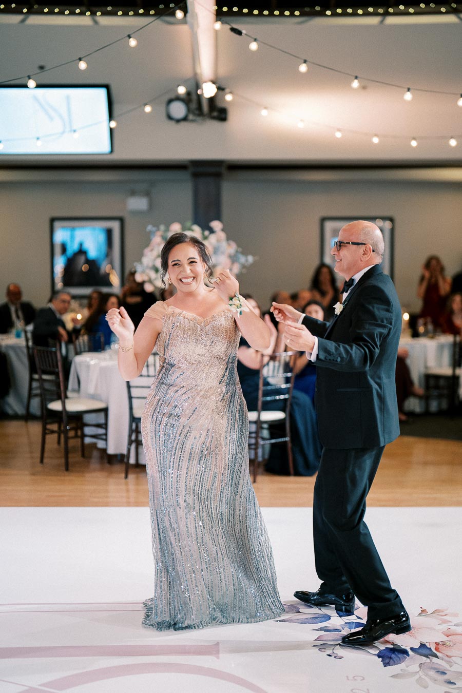 A joyful couple dancing at a wedding reception, surrounded by elegantly decorated tables and ambient string lights.