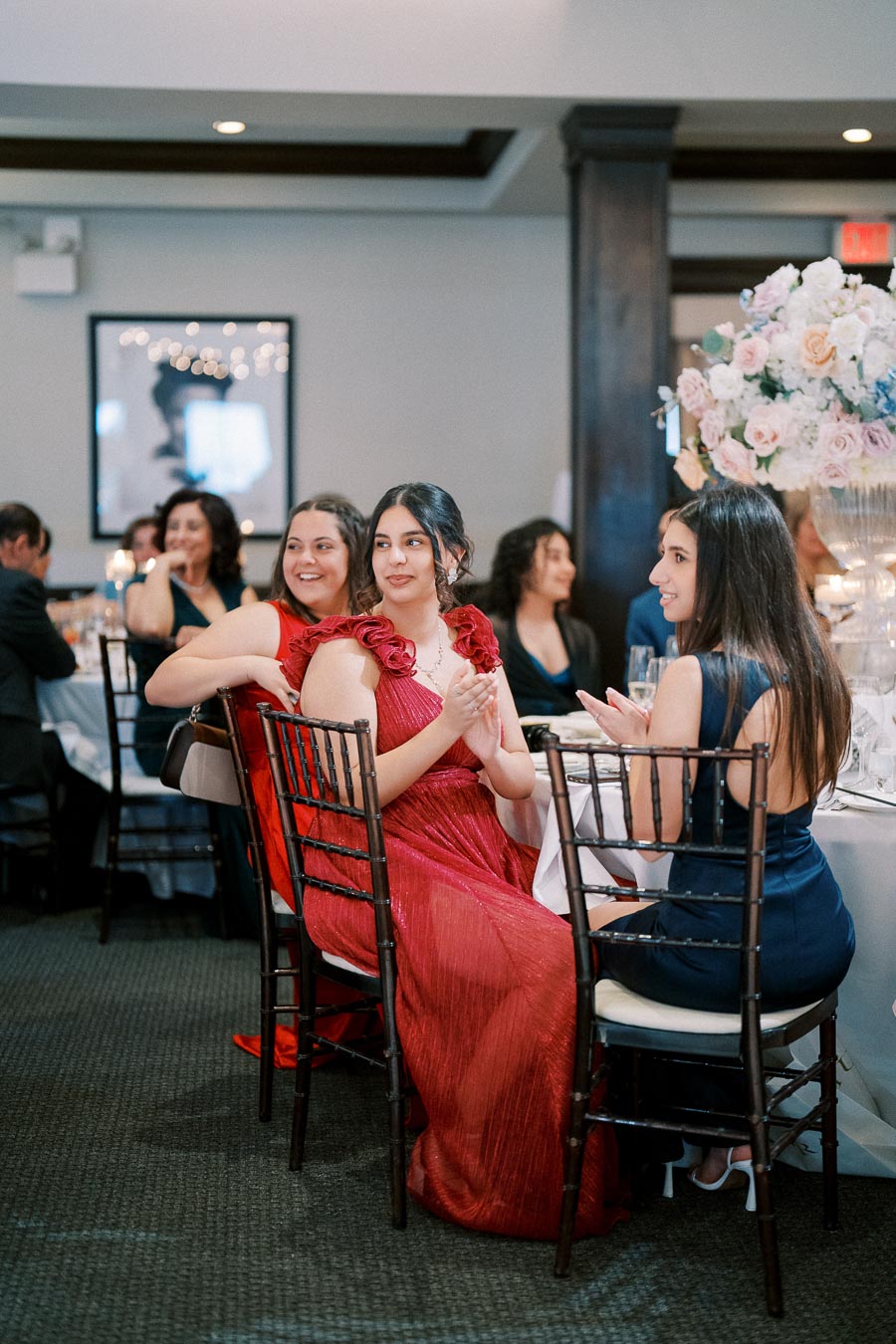 Three women in elegant dresses sitting at a formal event table, smiling and clapping, surrounded by other guests and a large floral centerpiece with pink and white roses.