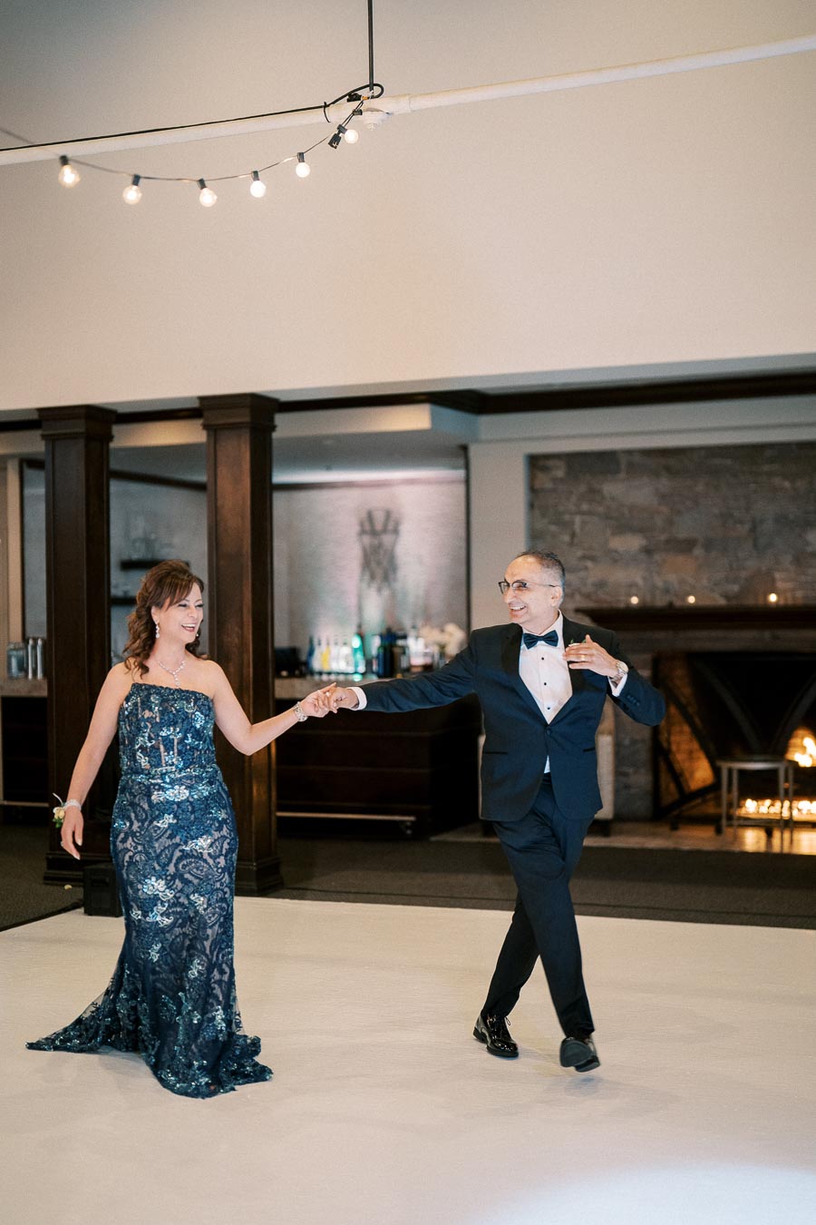 A couple elegantly dancing in formal attire under string lights at a sophisticated indoor event with a fireplace in the background.