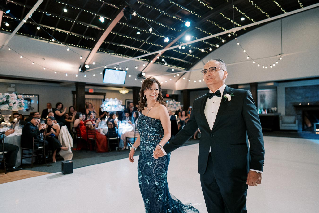 A couple elegantly dressed in formal attire shares a joyful moment on the dance floor during a wedding reception, surrounded by guests seated at decorated tables under twinkling string lights in a spacious venue.