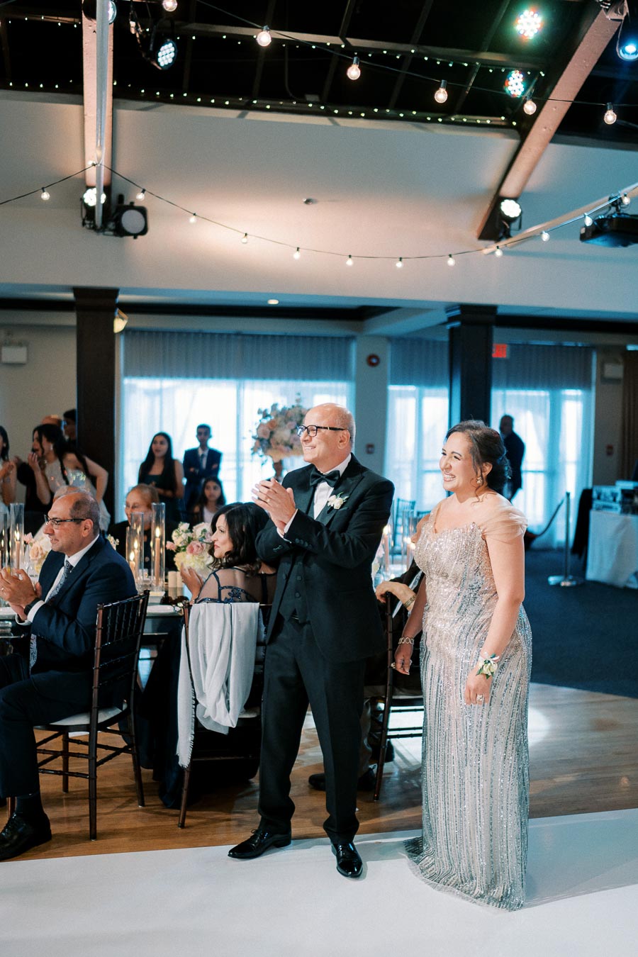Elegantly dressed couple clapping during a formal event with guests seated at decorated tables in a well-lit venue.