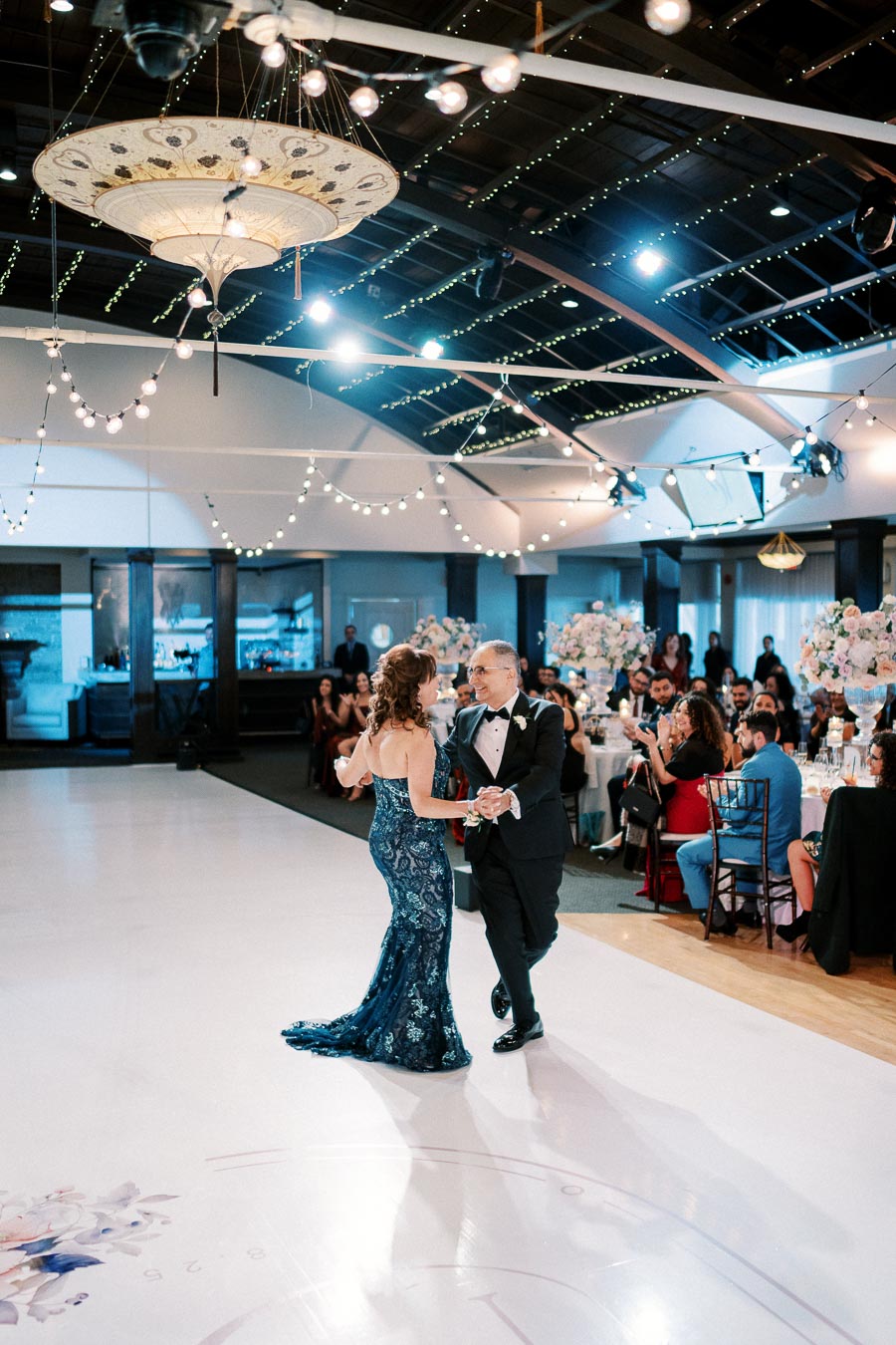 Elegant ballroom with string lights, featuring a couple dancing in formal attire at a wedding reception, surrounded by guests and floral centerpieces.