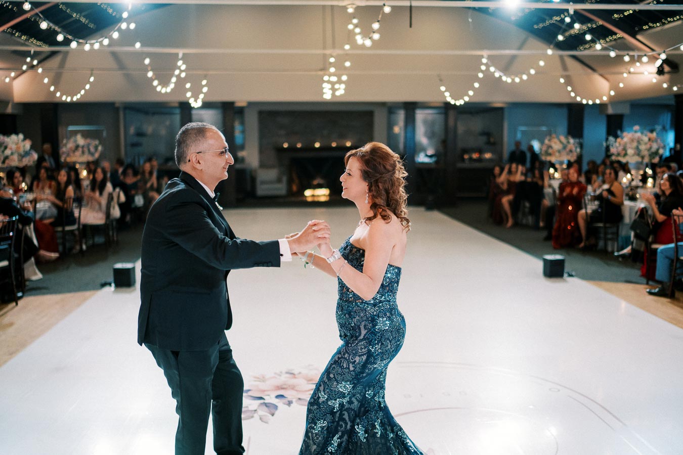 A couple elegantly dancing at a wedding reception in a beautifully decorated venue with string lights and guests in the background.