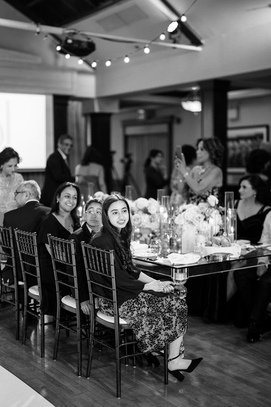 A black and white photo of a stylish indoor event with guests seated at a long table adorned with floral arrangements and candles. The room is softly lit, creating an elegant atmosphere. People are dressed in formal attire, and one individual is capturing the moment on a smartphone.