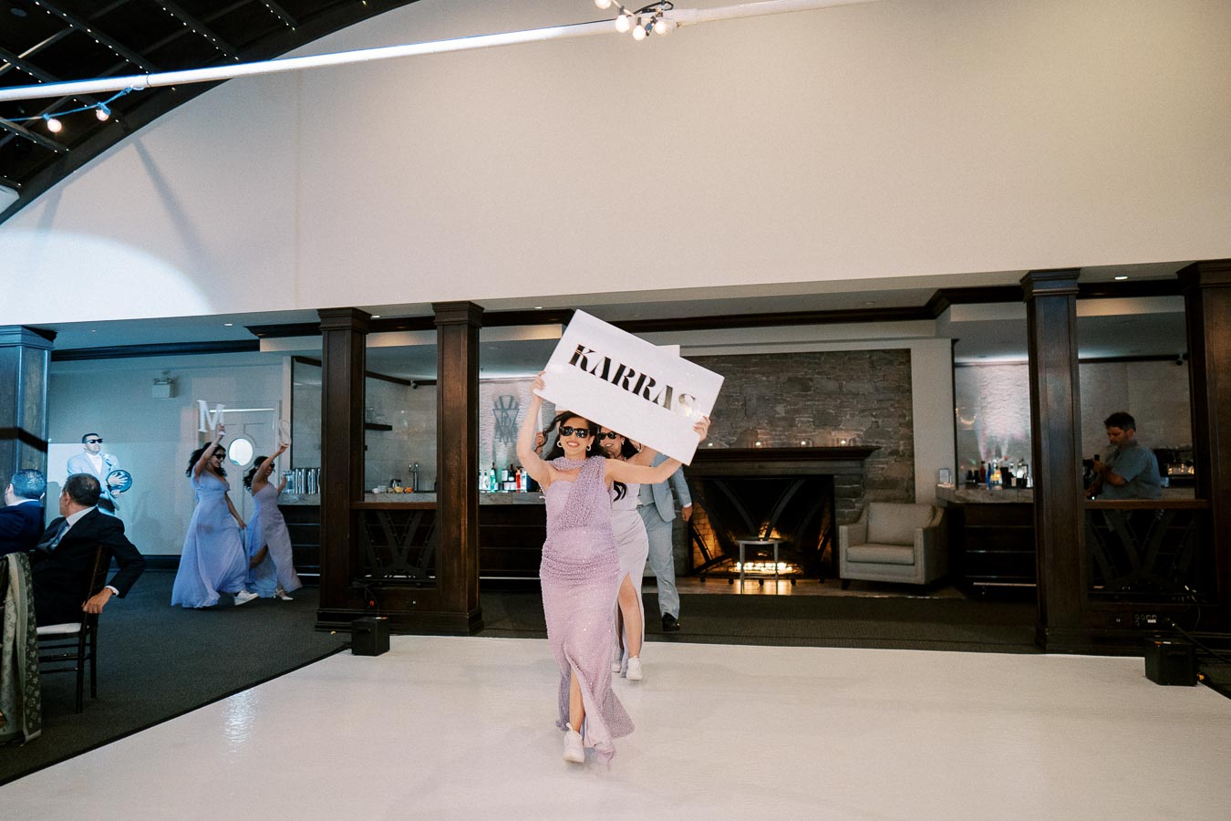 Guests in formal attire celebrate at a wedding reception, with one person holding a large sign that reads Karras, at a stylish indoor venue with a bar and stone fireplace in the background.