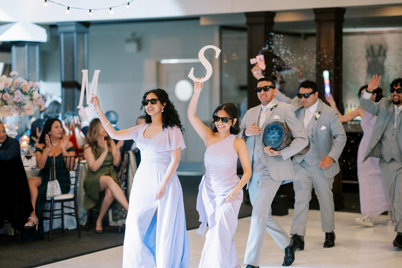 A group of well-dressed wedding party members in lavender attire and sunglasses energetically dance and hold up large letters inside a decorated reception venue.