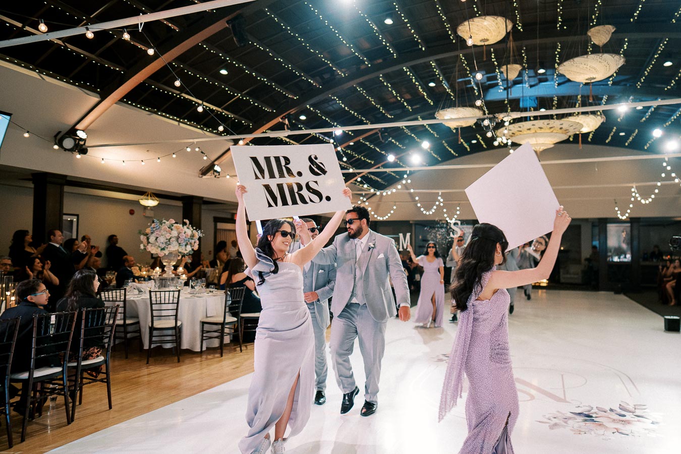 Guests cheer as a couple in stylish attire and sunglasses joyfully enter a wedding venue, holding a Mr. & Mrs. sign. The room is elegantly decorated with string lights and floral arrangements, enhancing the celebratory atmosphere.