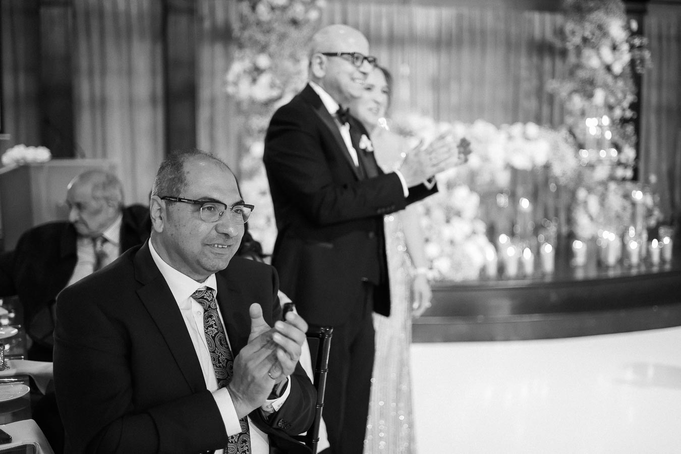 Black and white photograph of a formal event featuring a man in glasses and a suit clapping in the foreground, with two people clapping in the background amidst elegant decor and floral arrangements.
