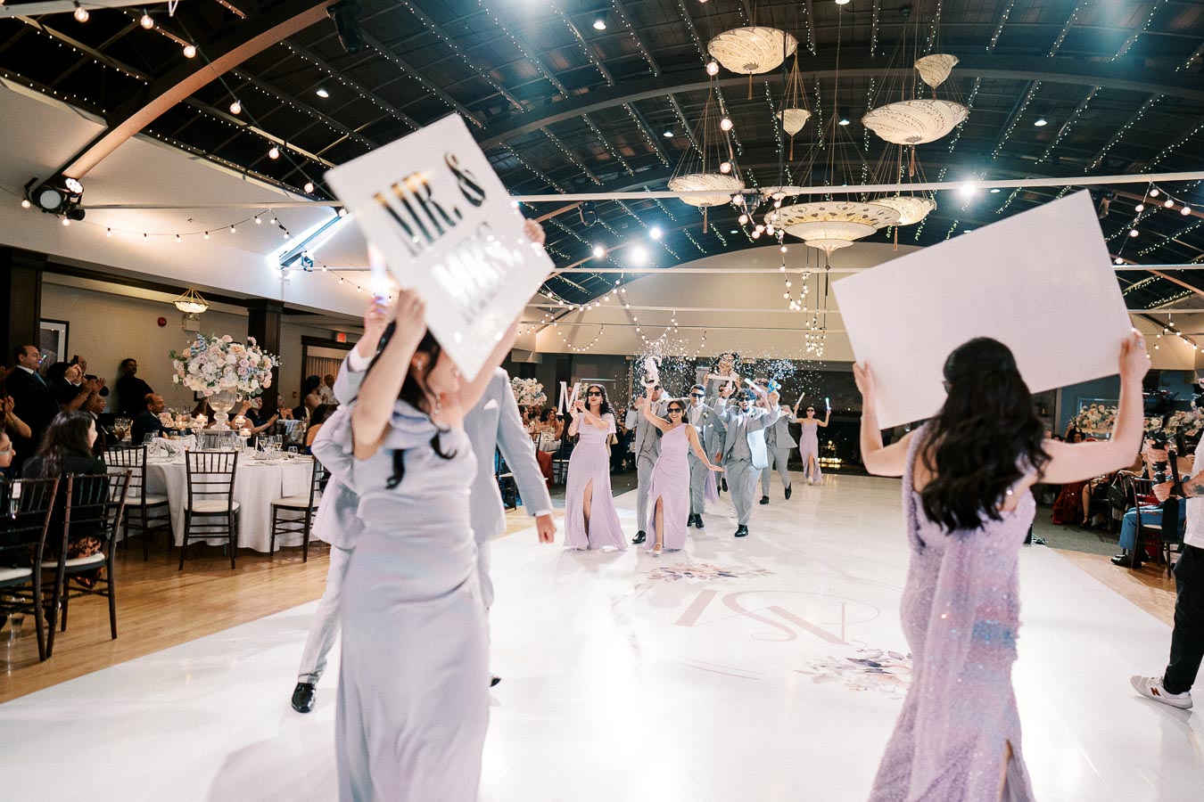 Wedding celebration with guests in formal attire dancing and holding signs on a decorated dance floor, surrounded by tables adorned with floral centerpieces and string lights in an elegant venue.