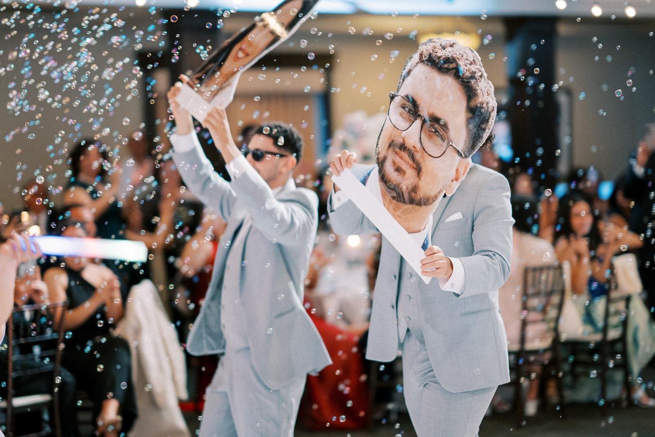 Two men in gray suits celebrating in a lively party setting, holding oversized head cutouts and surrounded by floating bubbles, creating a festive atmosphere with a cheering audience in the background.