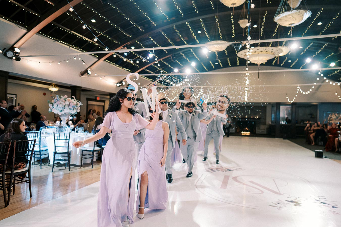 Wedding party procession with bridesmaids in lavender dresses and groomsmen in gray suits wearing sunglasses, under twinkling fairy lights in an elegant reception hall, creating a festive atmosphere.