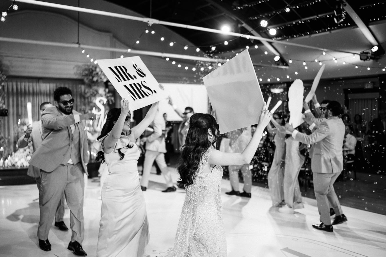 A lively wedding celebration with joyous guests dancing and holding up Mr. & Mrs. signs, captured in black and white.