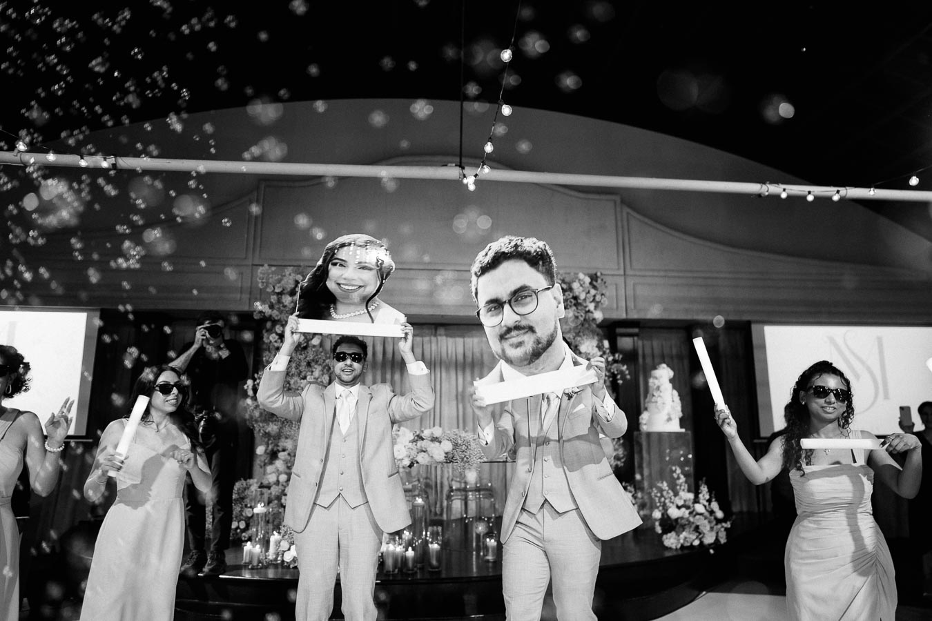 A group of people dressed in formal attire, holding oversized face cutouts, celebrating at a wedding reception with bubbles floating around, creating a joyful and lively atmosphere.