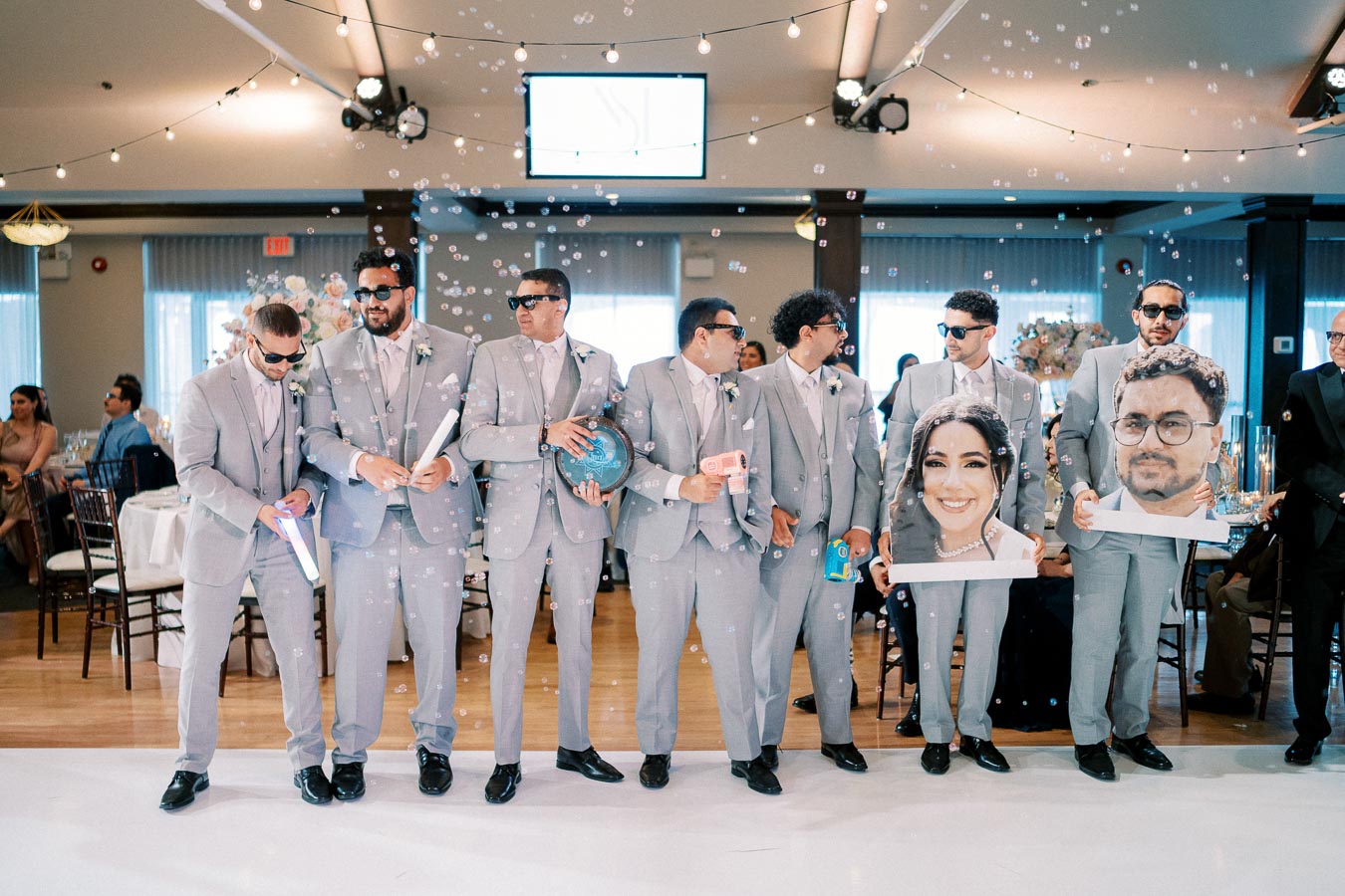 A group of groomsmen in matching gray suits and sunglasses playfully holding bubble guns and large face cutouts at a wedding reception, with bubble decorations and string lights illuminating the venue.