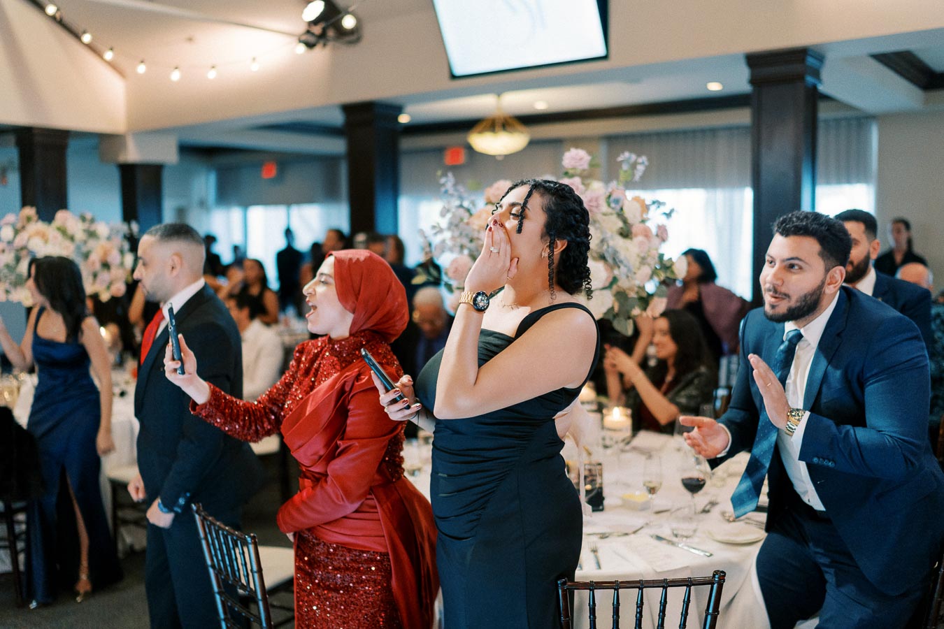 Guests excitedly cheering and capturing moments on their phones at a lively wedding reception, with elegant decor and floral arrangements in the background.