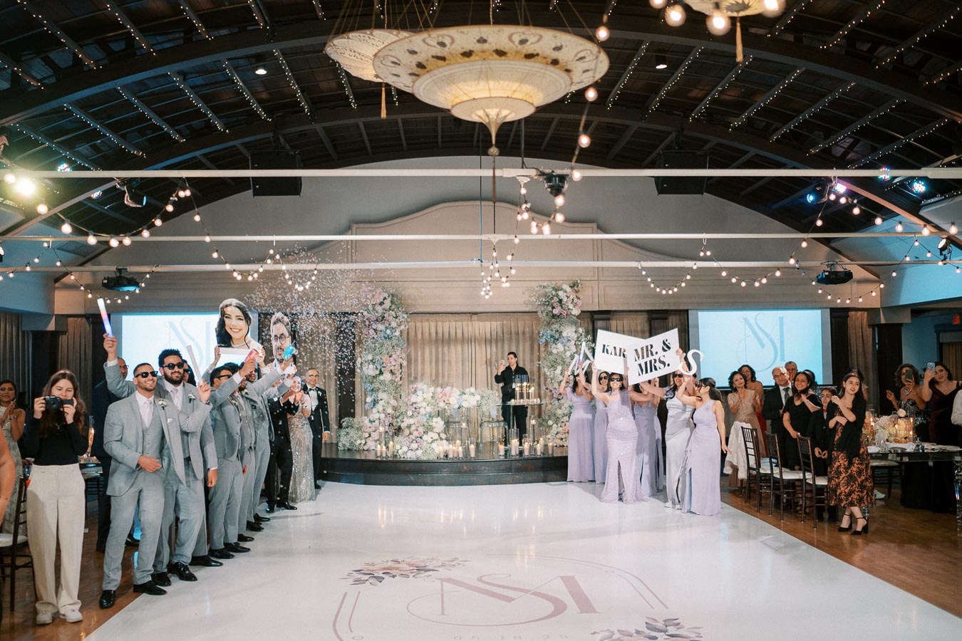 Wedding reception in a beautifully decorated hall with guests celebrating; people in suits and dresses hold signs and props, surrounded by floral arrangements and string lights.