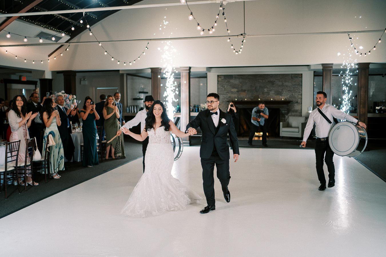A newlywed couple joyfully enters their wedding reception hand in hand, surrounded by drummers and applauding guests, under twinkling string lights and sparkling effects in an elegant venue.
