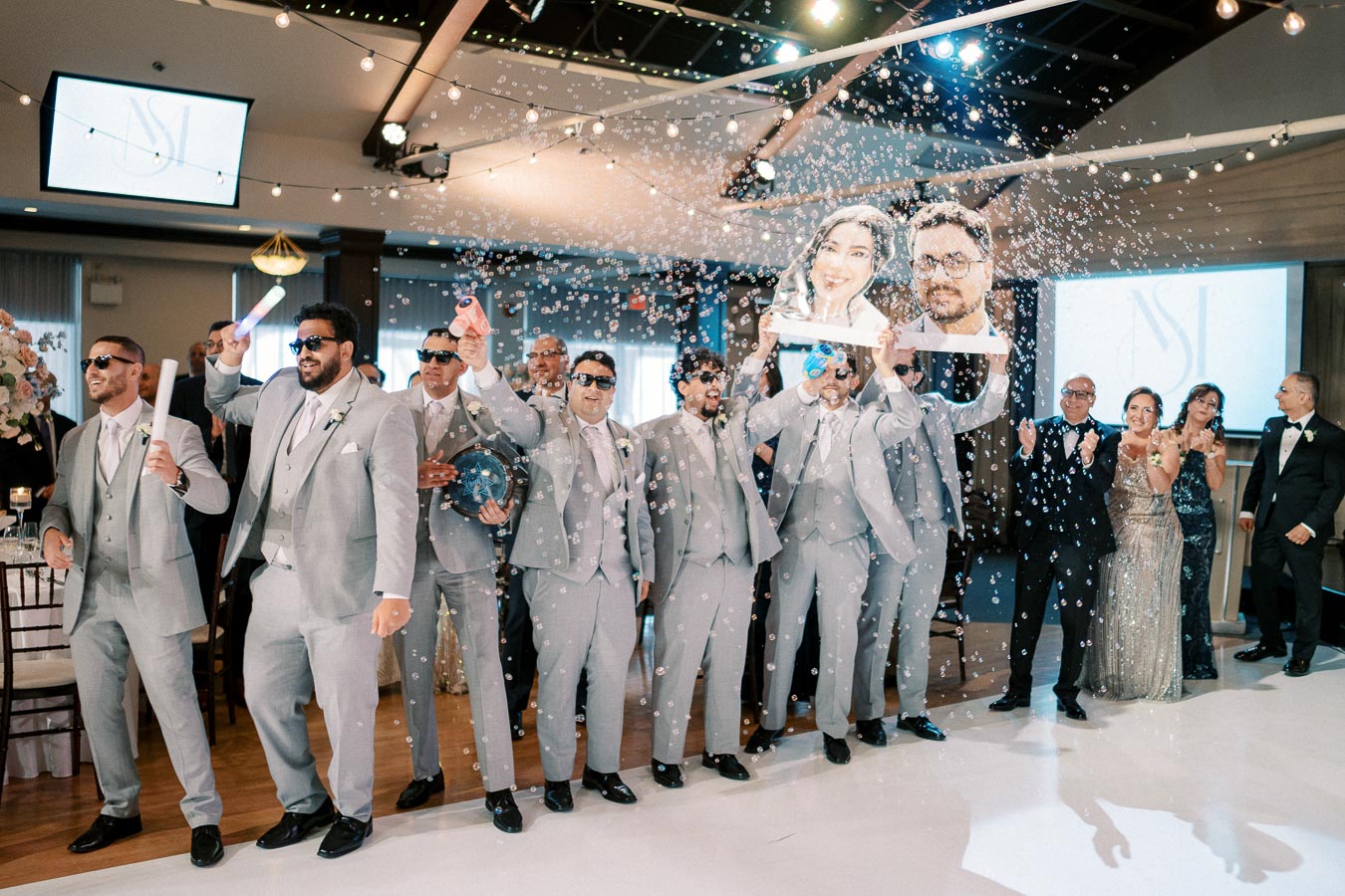 Group of groomsmen in matching gray suits and sunglasses celebrating energetically with bubbles at a wedding reception, holding large cut-out photos of the couple, surrounded by joyful guests and festive decor.