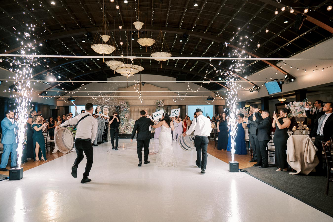 Wedding celebration with a bride and groom dancing on a white floor, surrounded by guests. Bright sparklers illuminate the venue, creating a festive atmosphere. Musicians with drums lead the procession, with joyful guests applauding and capturing the moment on their phones. Elegant lighting and floral decorations enhance the sophisticated setting.