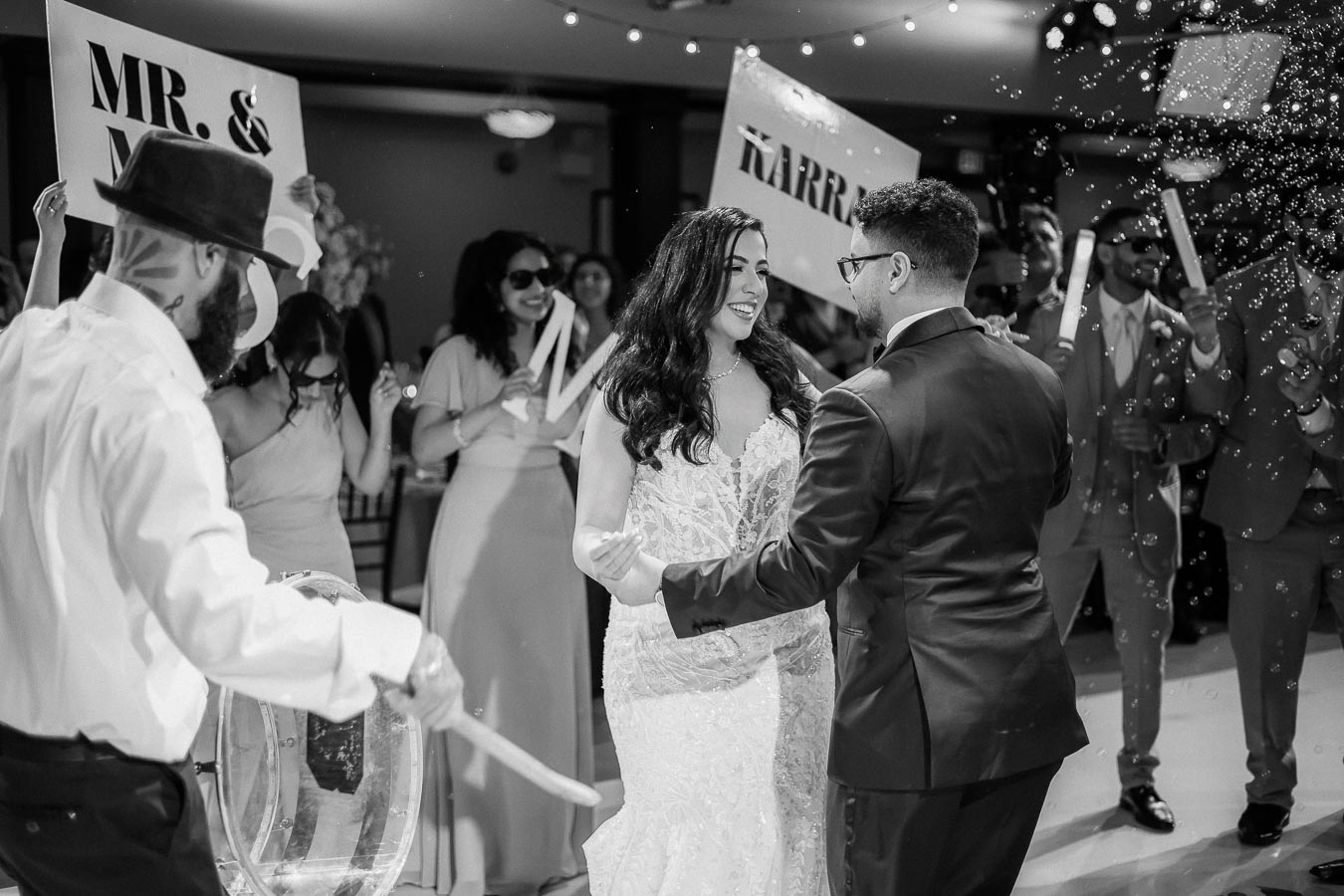 A bride and groom share a dance at their wedding reception, surrounded by smiling guests holding celebratory signs. Bubbles float in the air, enhancing the joyful atmosphere.