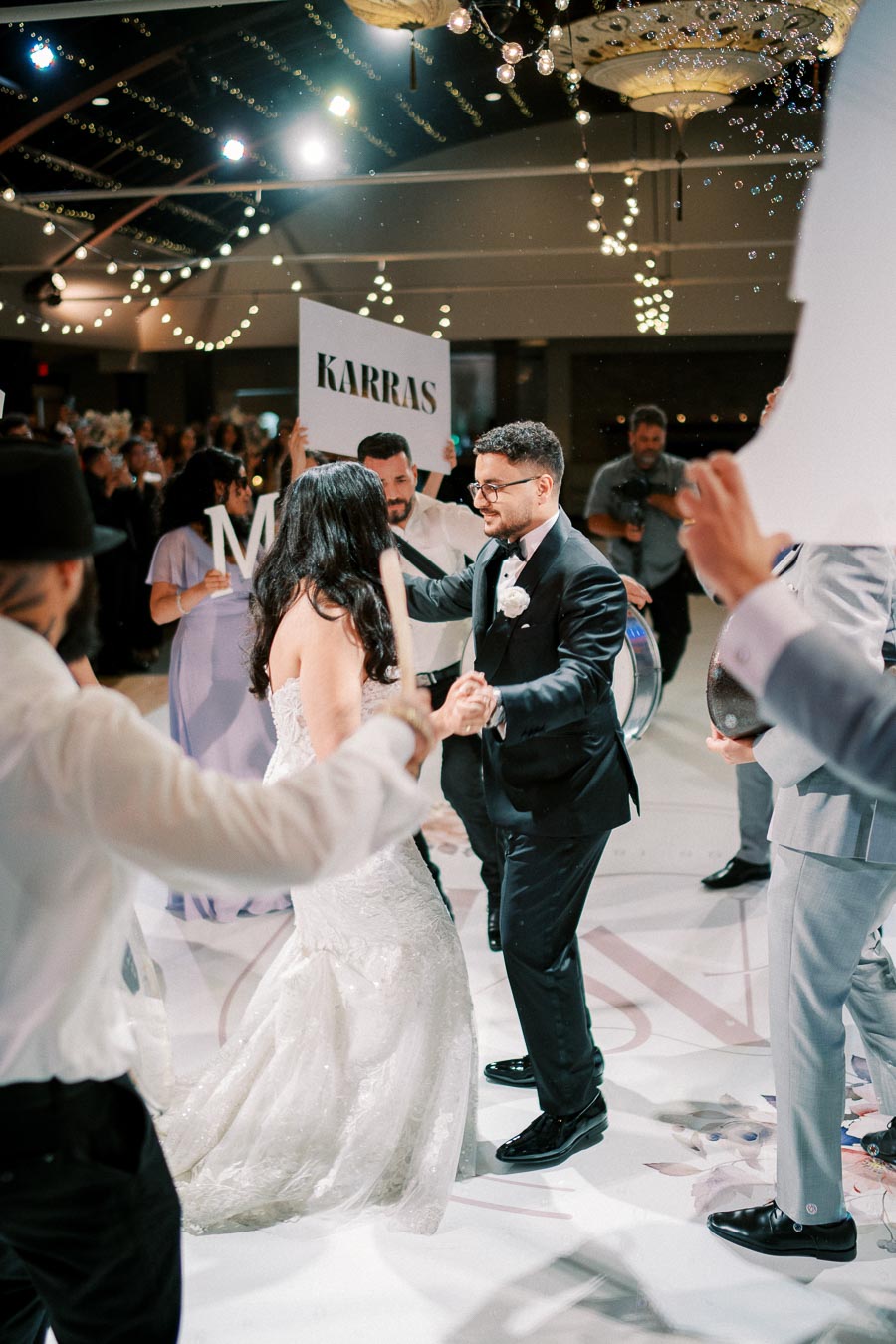 A joyful wedding celebration with a bride and groom dancing surrounded by guests at a reception venue, decorated with string lights and warm ambiance.