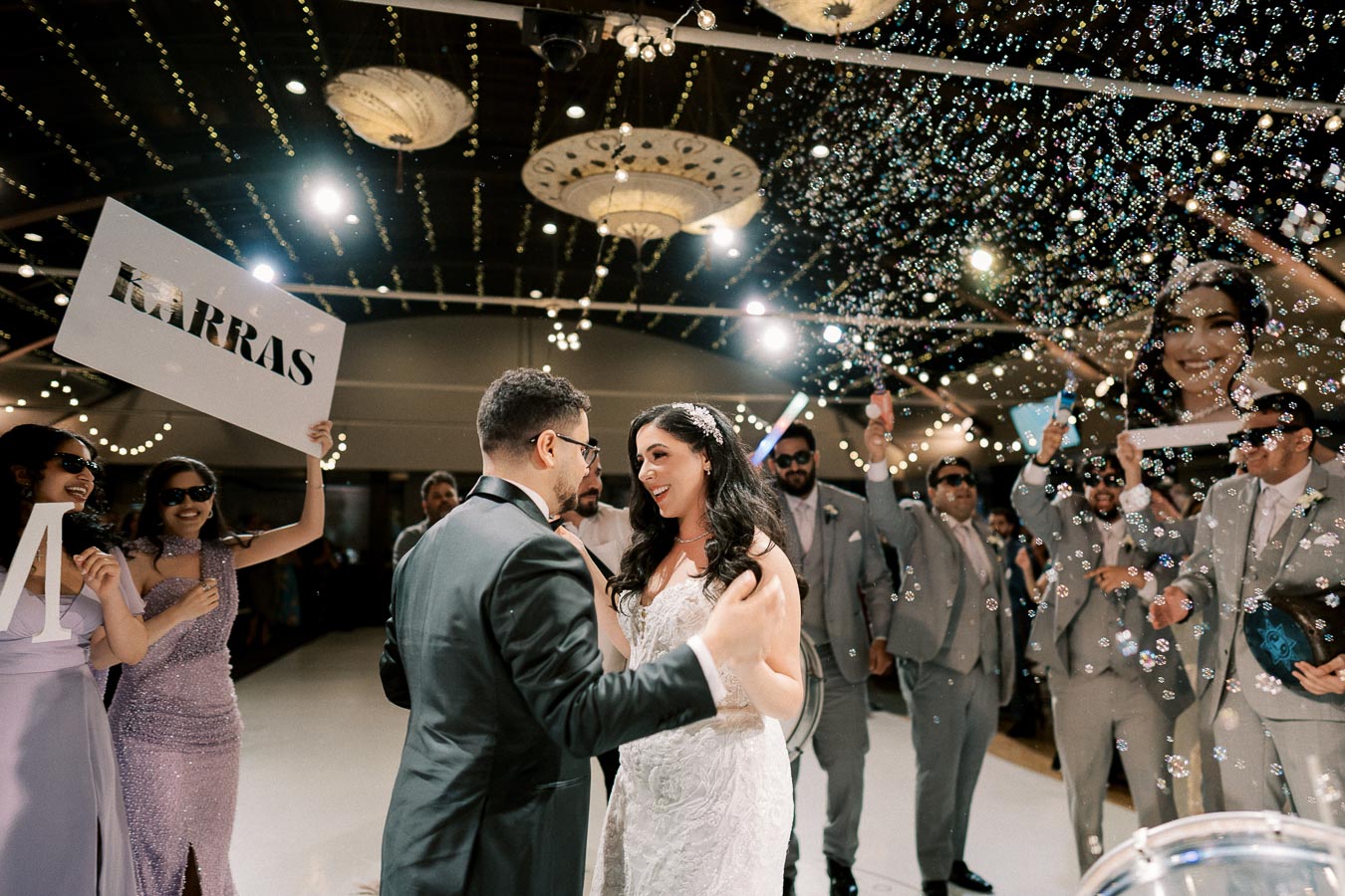 A bride and groom share a dance on their wedding day surrounded by joyful guests celebrating with bubbles under twinkling lights in an elegantly decorated venue.
