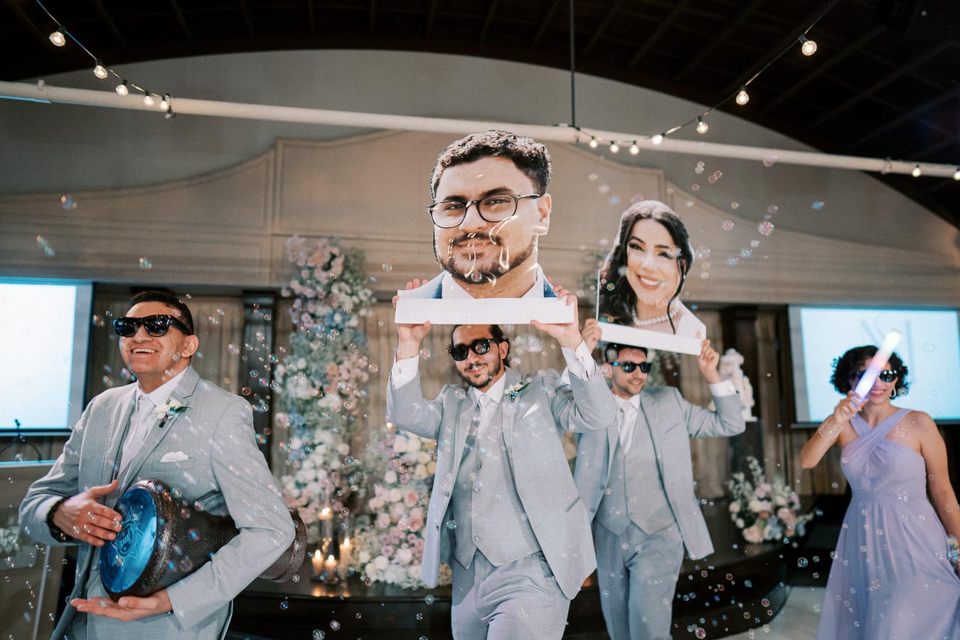 Wedding celebration with groomsmen in light gray suits and sunglasses holding large face cutouts, surrounded by bubbles and festive decor.