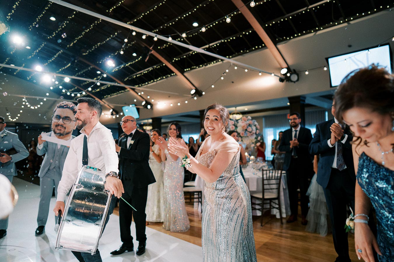 A festive wedding reception with guests clapping and smiling, as a man plays a drum. The venue is decorated with string lights, creating a joyful atmosphere.