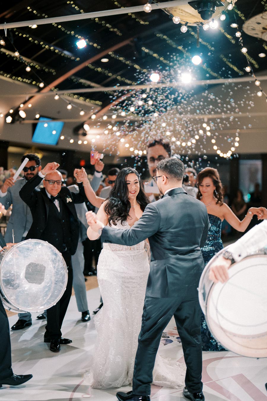 A bride and groom dance joyfully at their wedding reception, surrounded by guests celebrating with music and confetti under festive string lights in an elegant venue.