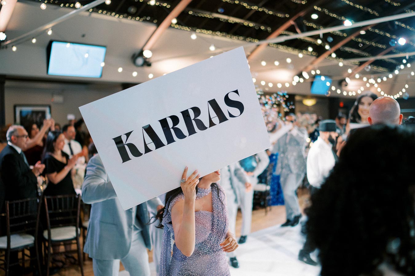 A person holding a large sign reading Karras during a festive indoor event with a crowd clapping in the background, string lights hanging from the ceiling.