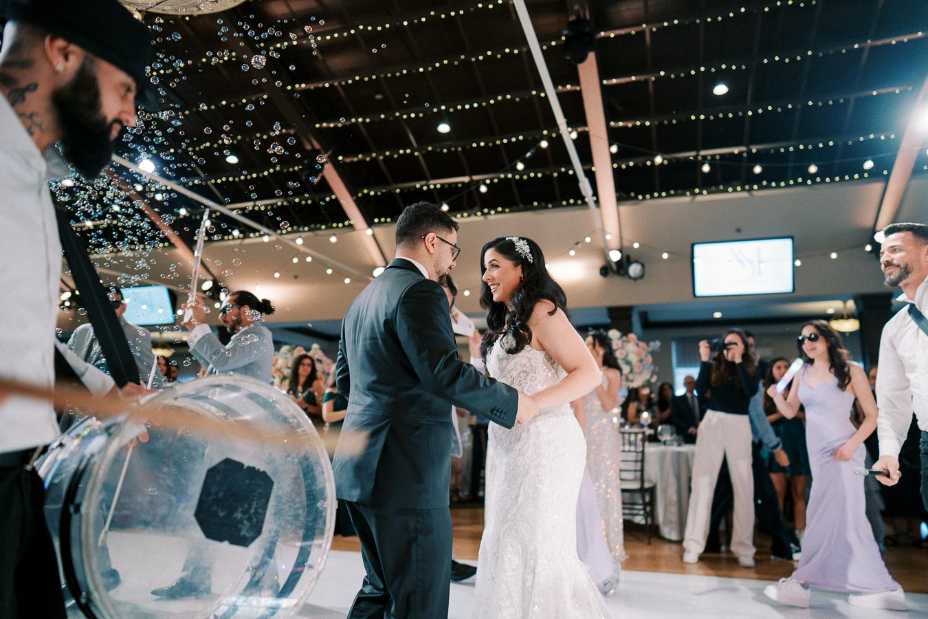 Bride and groom dancing joyfully on their wedding day surrounded by guests and a lively band, with twinkling lights and bubbles enhancing the celebratory atmosphere in the venue.
