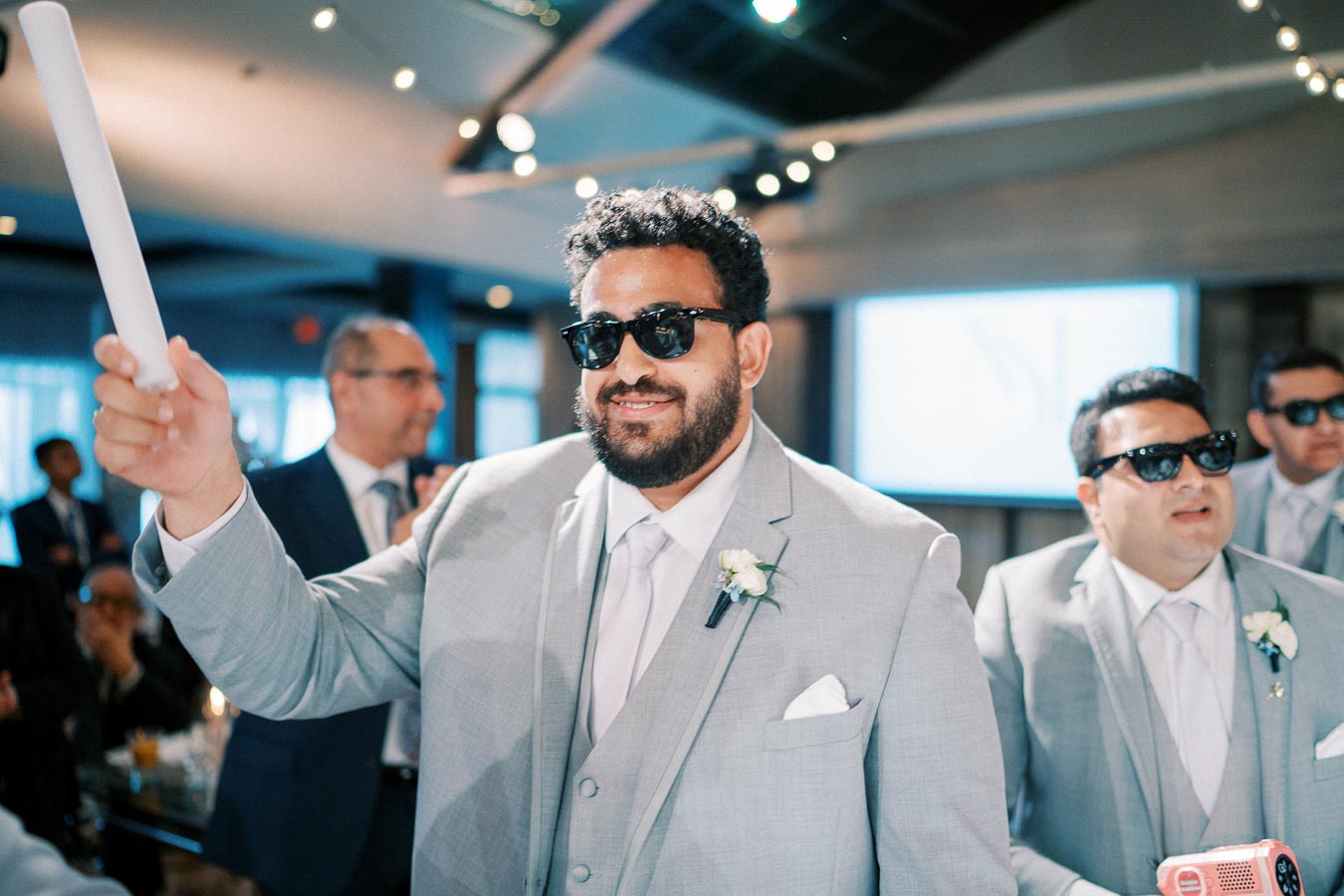Groomsman wearing sunglasses and a light gray suit holding a rolled-up paper at a wedding reception.