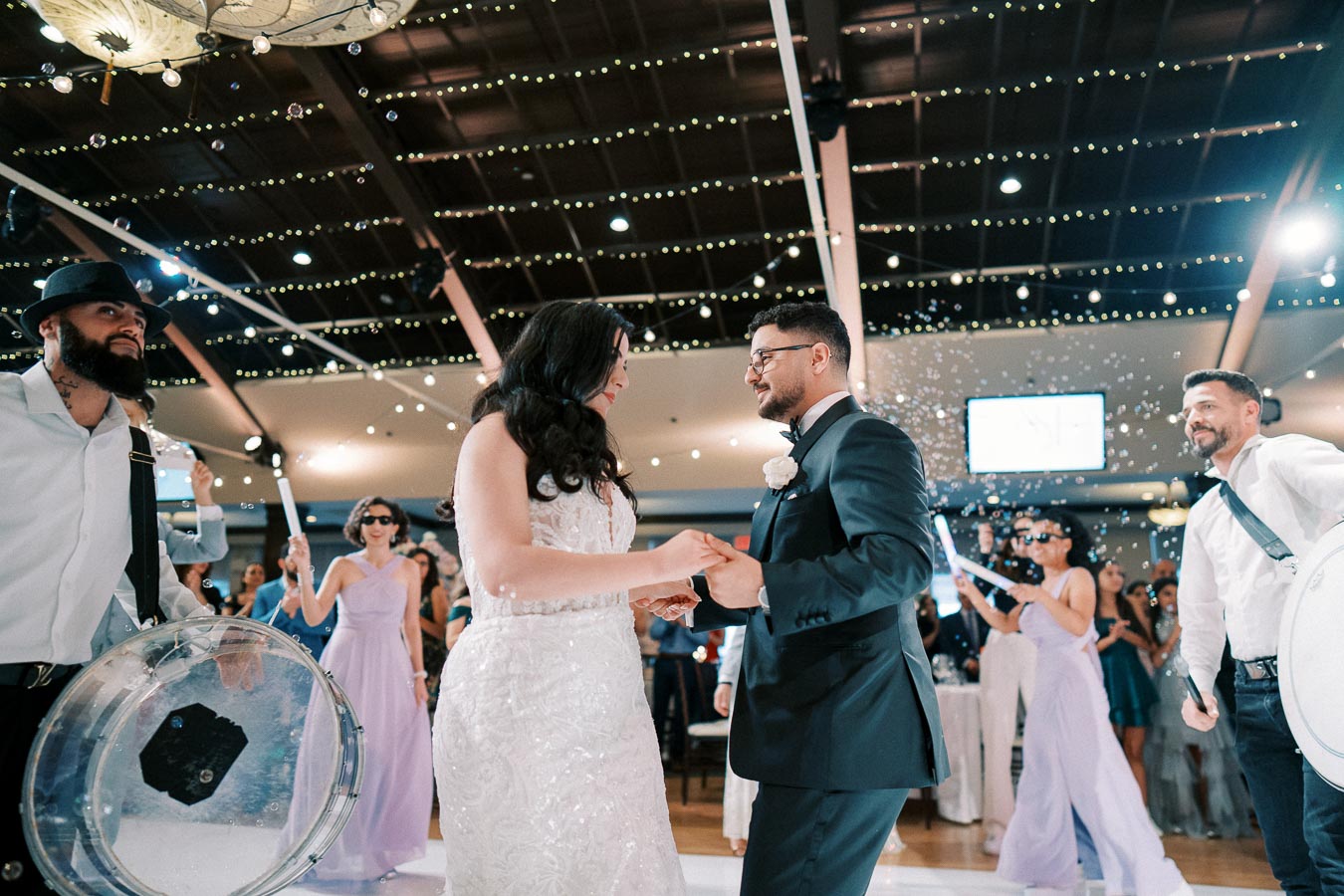 A bride and groom hold hands while dancing at a wedding reception, surrounded by guests in elegant attire. The venue is decorated with twinkling lights, and musicians play in the background, adding to the festive atmosphere.