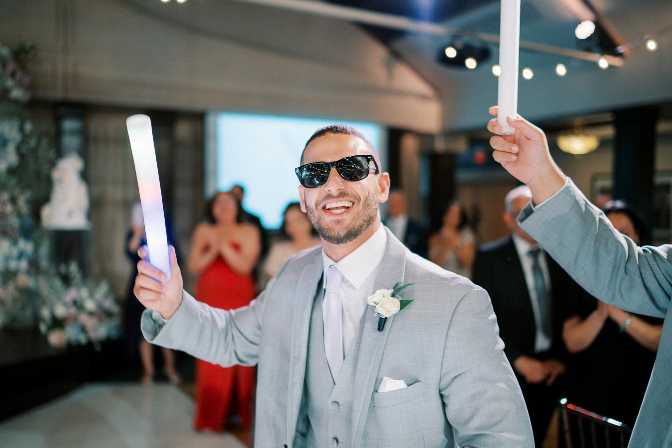 A joyful man in a light gray suit and sunglasses holding a glowing light stick at an indoor celebration or wedding, surrounded by smiling guests and festive decorations.