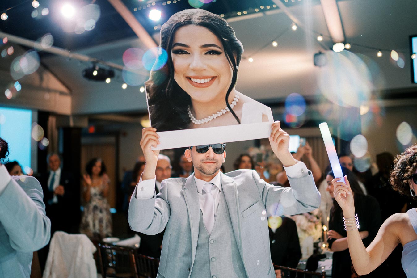 Man in a light gray suit and sunglasses holding a large smiling face cutout at a lively wedding reception, surrounded by guests under twinkling lights.