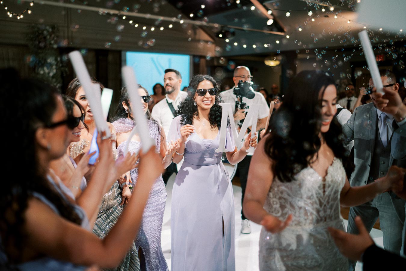 A group of people celebrating at a wedding reception, with guests wearing formal attire and sunglasses. A woman in a lavender dress is holding a letter M, surrounded by bubbles and festive lighting.