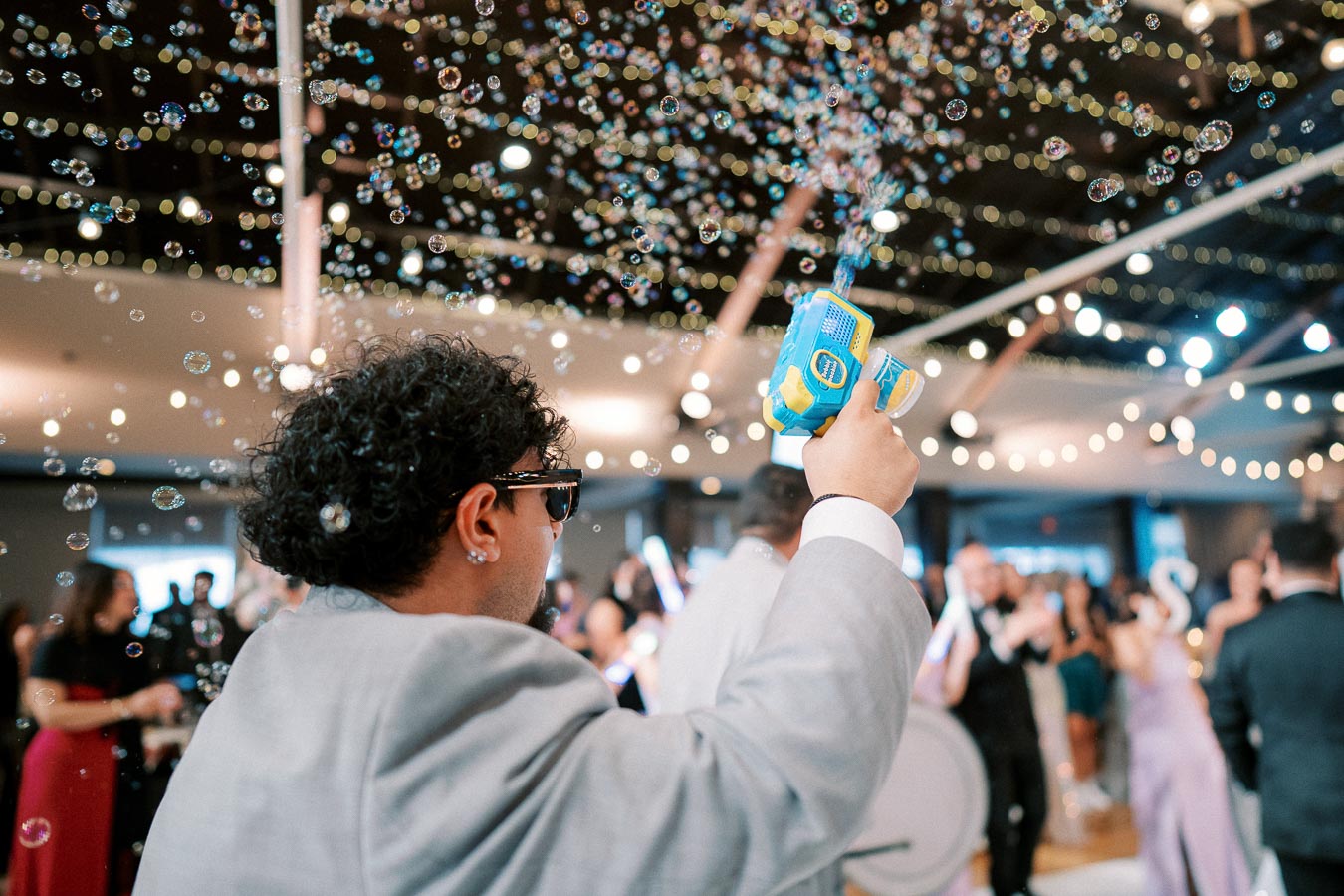 Man in a gray suit using a bubble gun at a lively indoor celebration, surrounded by floating bubbles and festive string lights.