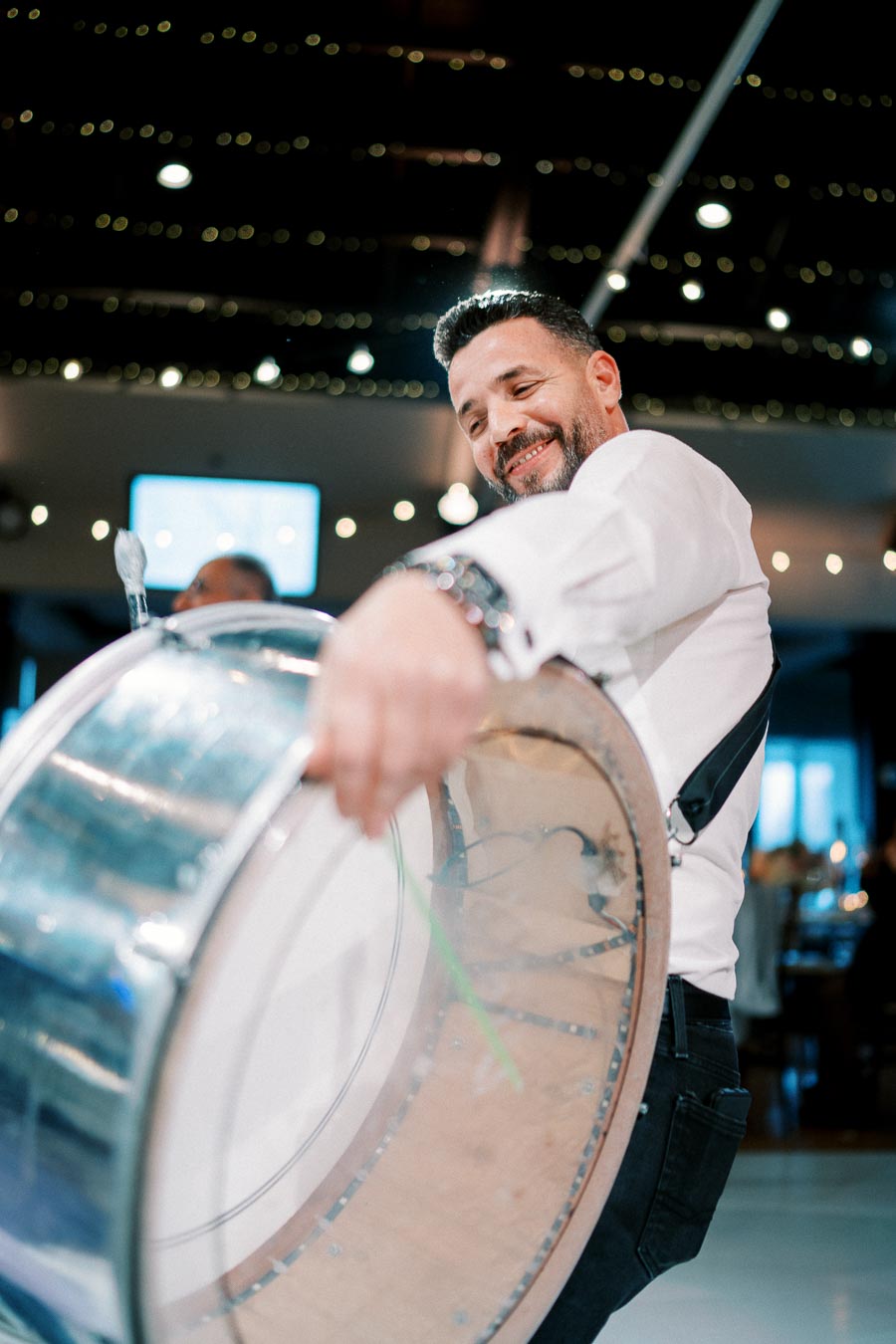 A joyful musician playing a large metallic drum in a festive indoor setting with twinkling lights in the background.