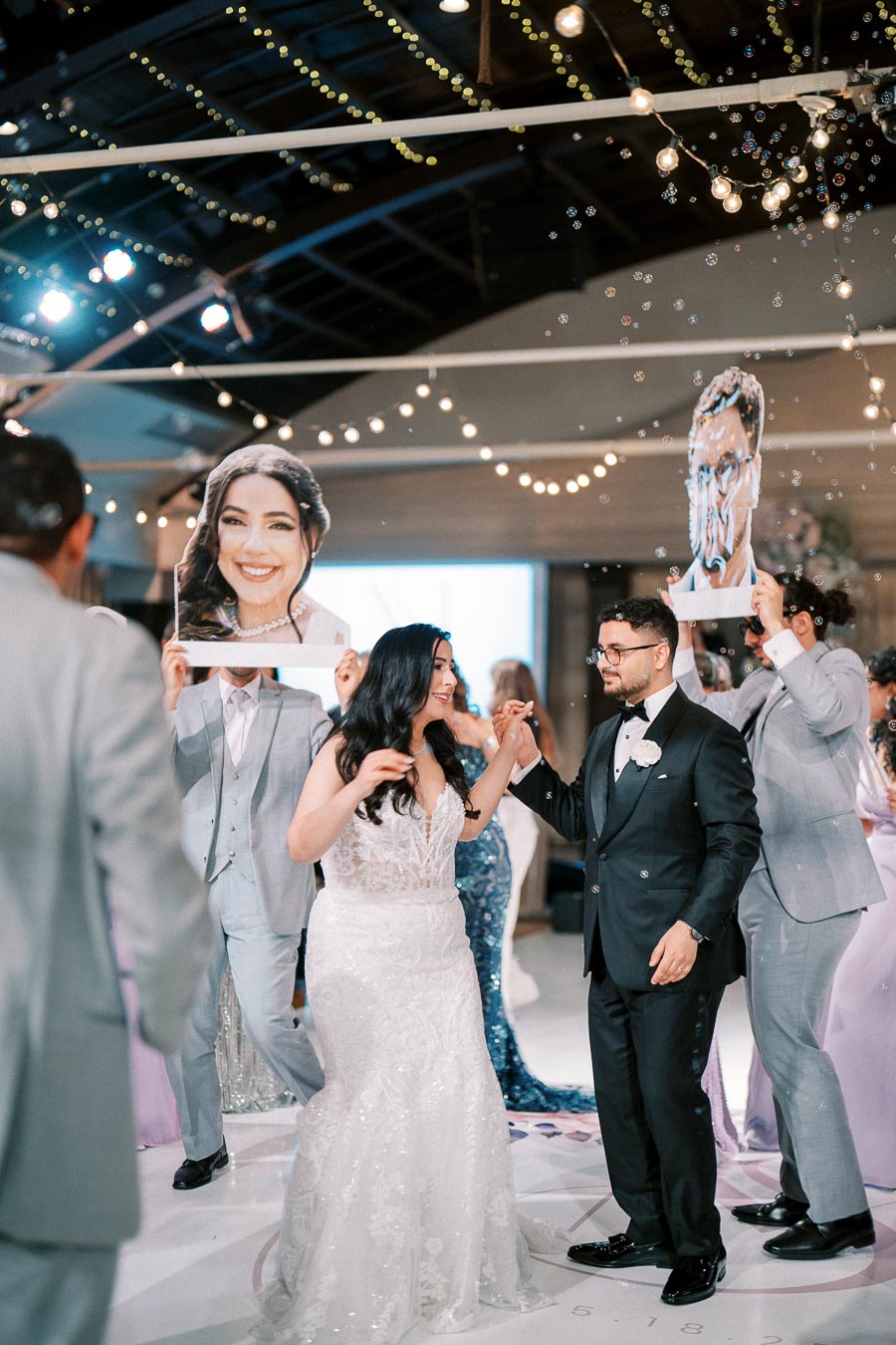 A bride and groom dance joyfully at their wedding reception surrounded by bubbles and guests holding large cutouts of their faces, with twinkling string lights decorating the venue.