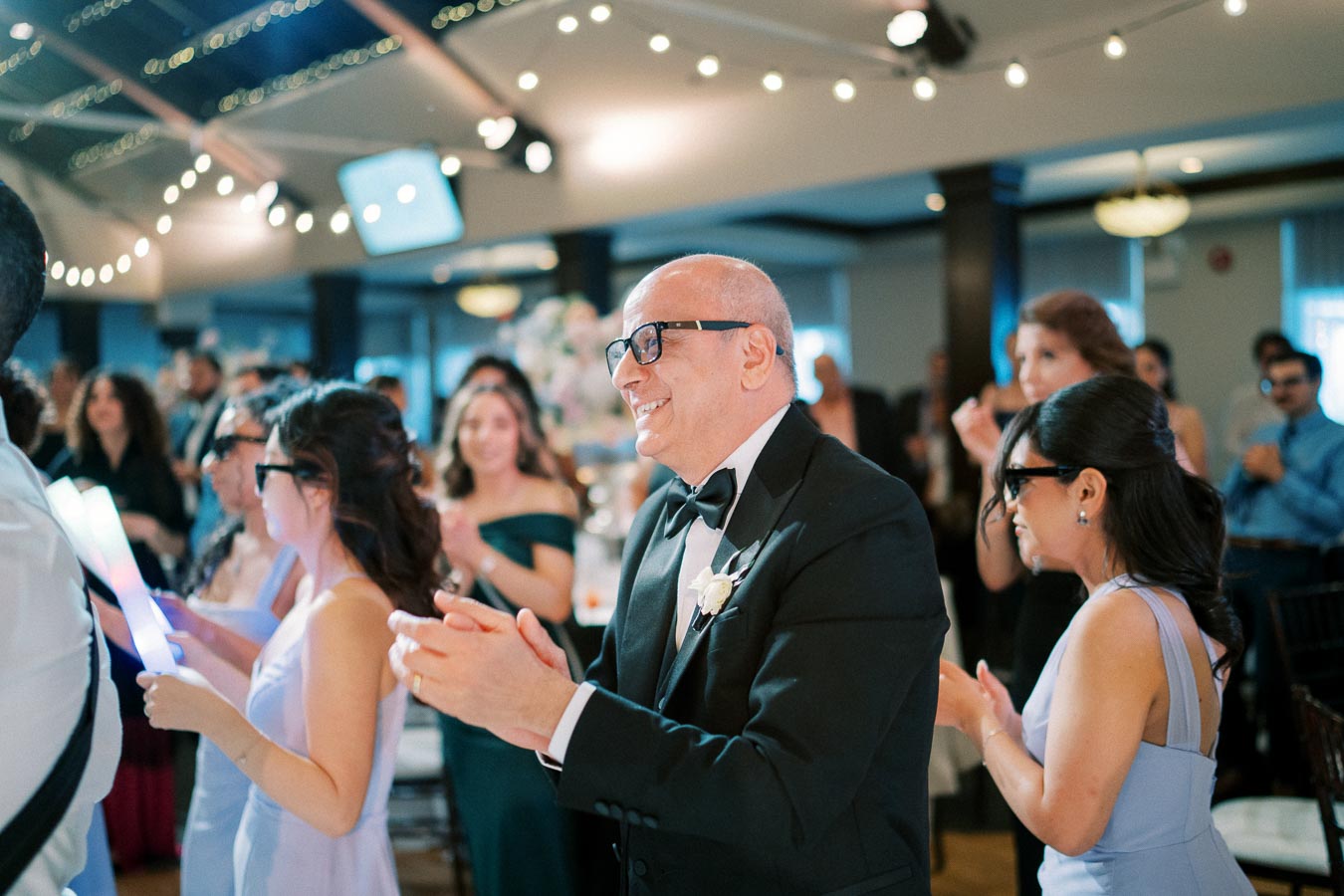 Elegant indoor wedding celebration with guests wearing formal attire, clapping and smiling under ambient string lights in a beautifully decorated venue.