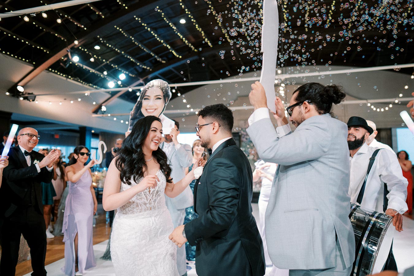 A joyful wedding reception with a bride and groom dancing among guests, festive lights, and bubbles in the background. Guests clap and celebrate, creating a lively atmosphere.