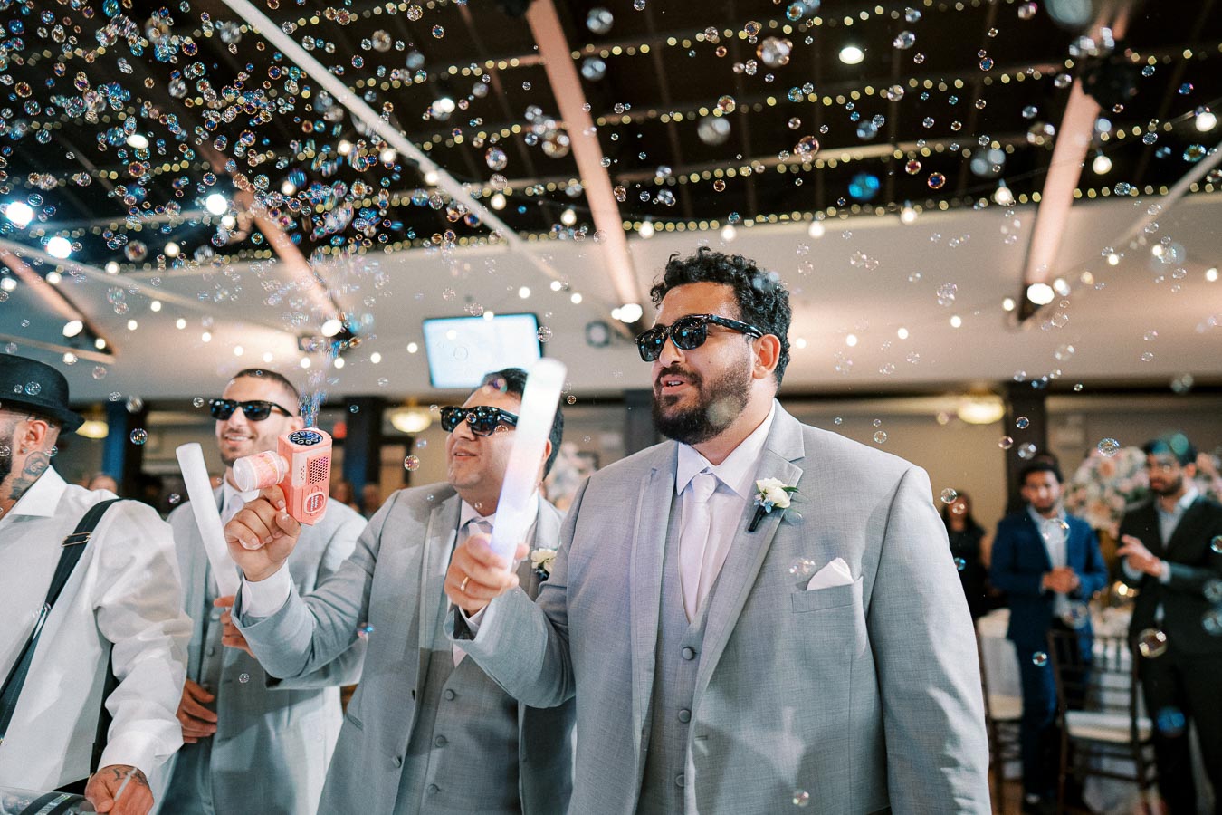 Groomsmen in gray suits and sunglasses enjoying a festive wedding reception with bubbles floating in the air.