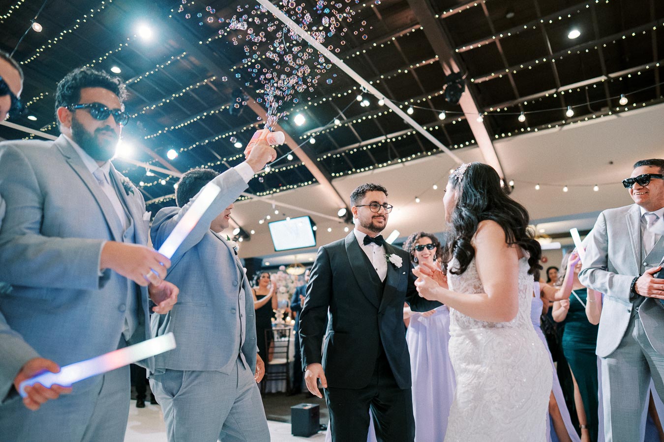 Wedding reception celebration with bride and groom dancing surrounded by guests in formal attire, holding light sticks and blowing bubbles in a festive, illuminated venue.