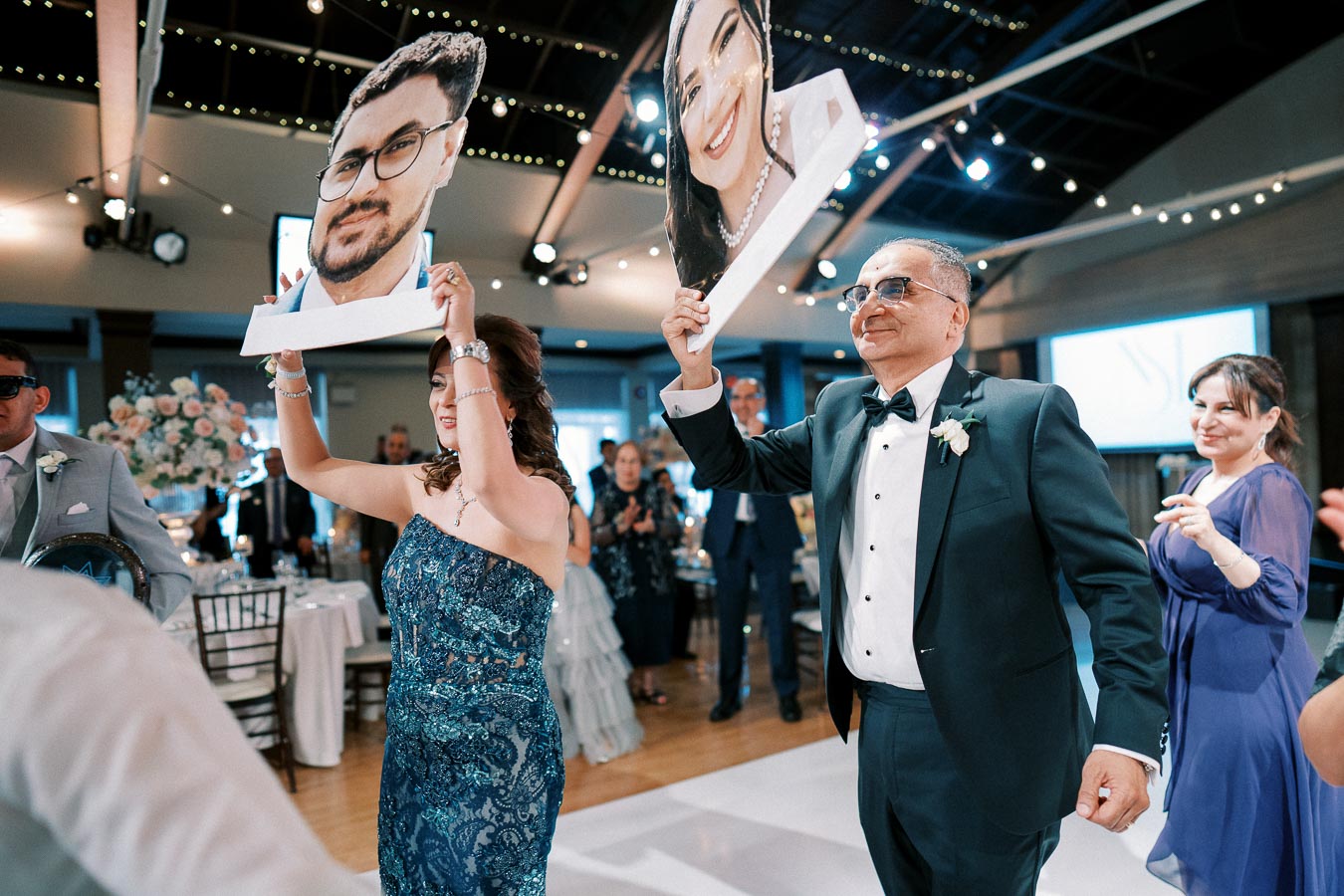 People celebrating at a wedding reception, holding large cutouts of a smiling bride and groom's faces, under string lights in a decorated venue.