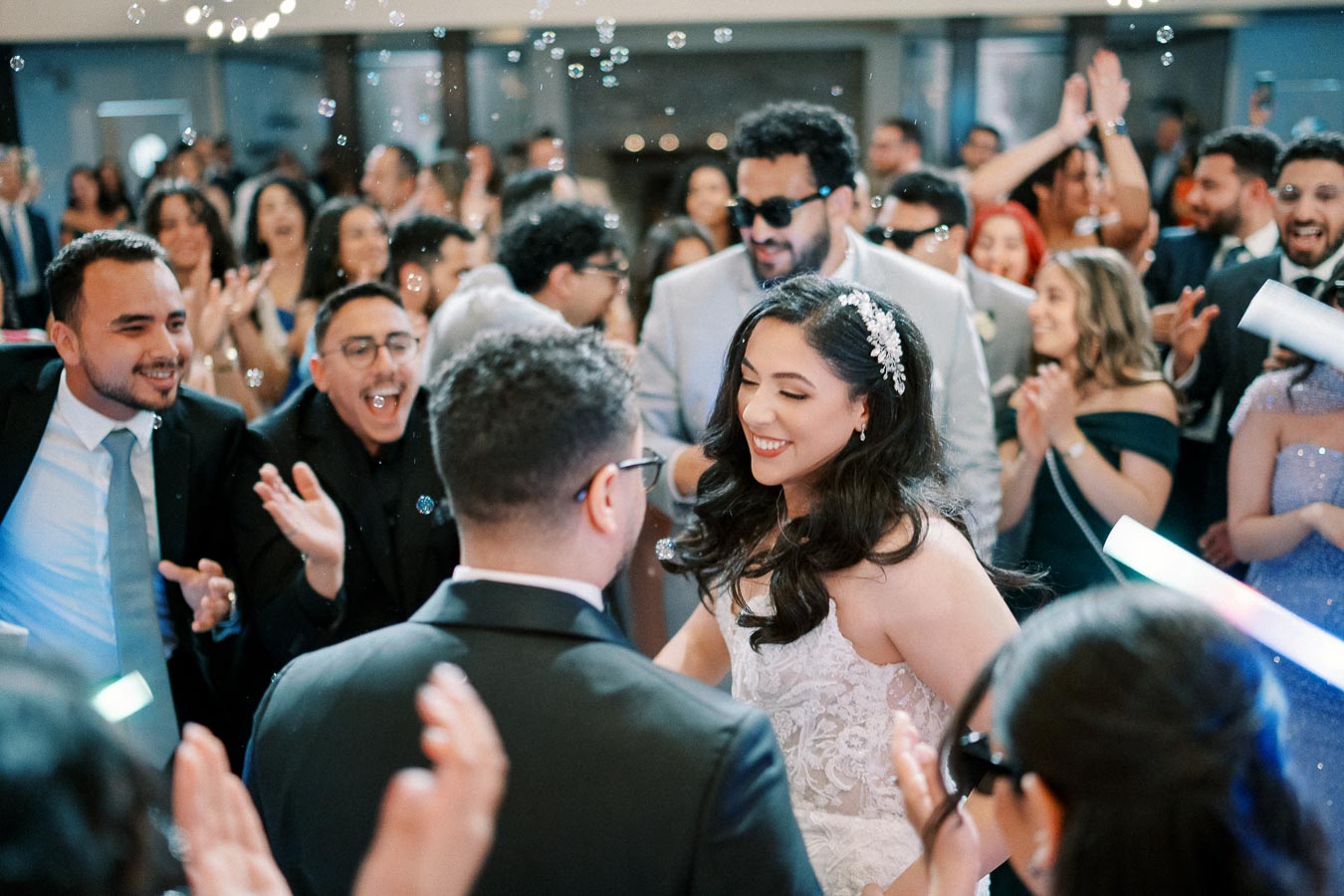 Group of people celebrating at a joyful wedding reception, with bride smiling and guests clapping and dancing, in a lively and festive atmosphere.