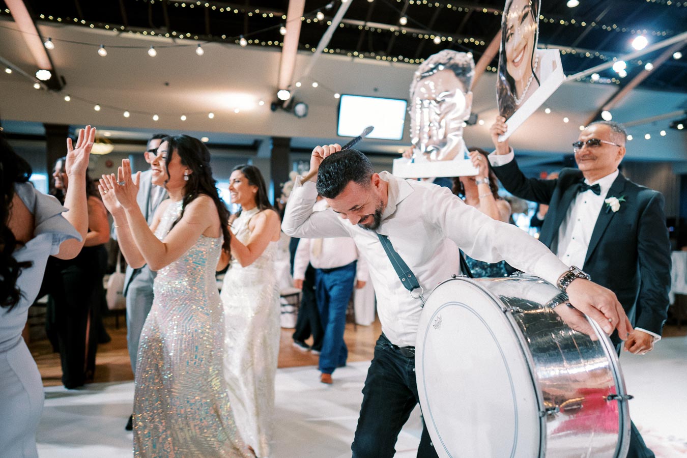 A lively wedding celebration with guests dancing joyfully, a man energetically playing a drum, and people holding large cutouts under twinkling lights in an elegant venue.